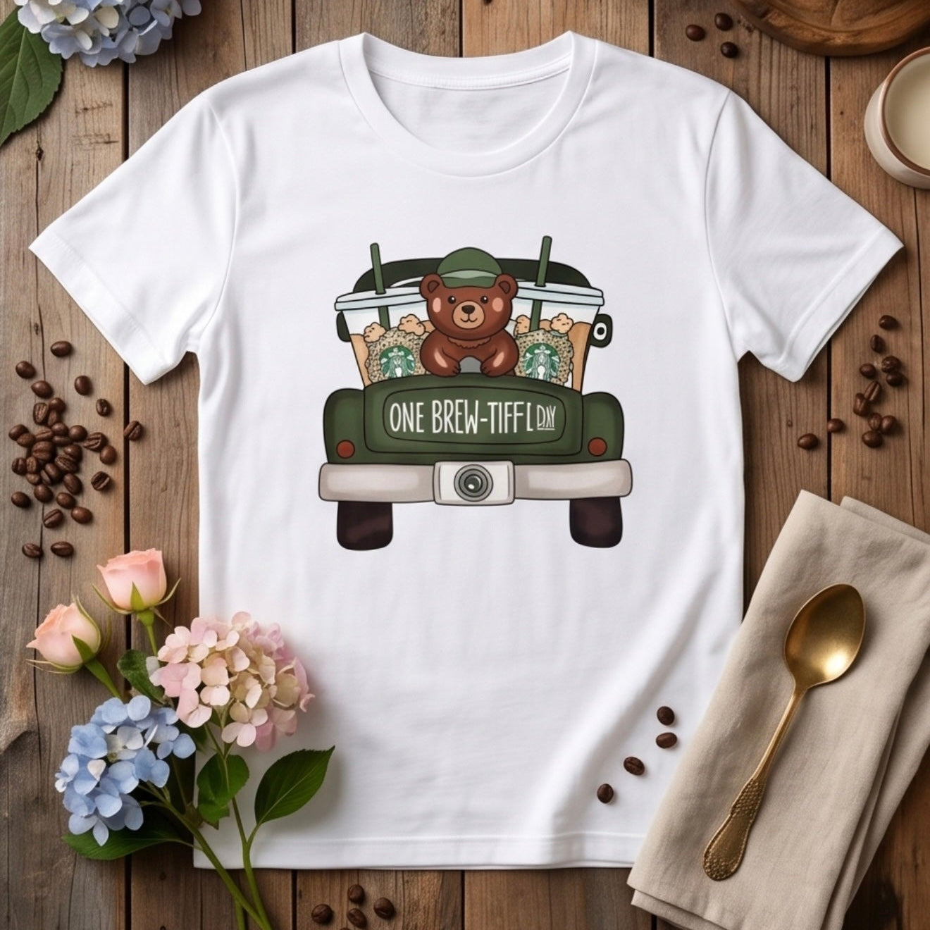 White t-shirt with a bear driving a truck graphic on a wooden surface with flowers and coffee beans.