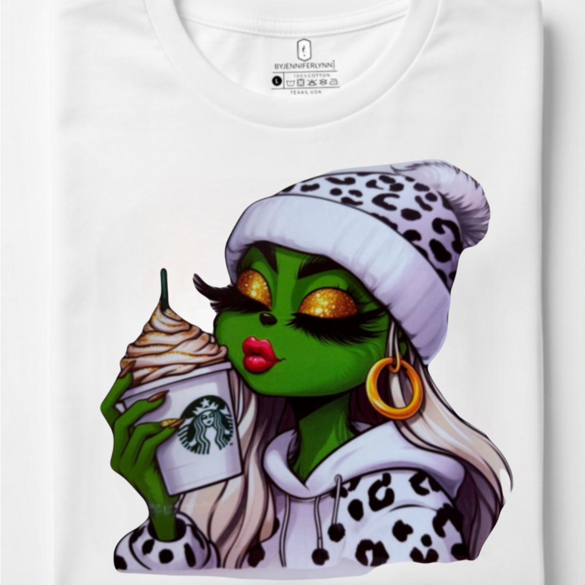 A folded white t-shirt with a colorful graphic print of a green-skinned woman with long white hair, wearing a leopard-print beanie and holding a Starbucks cup.
