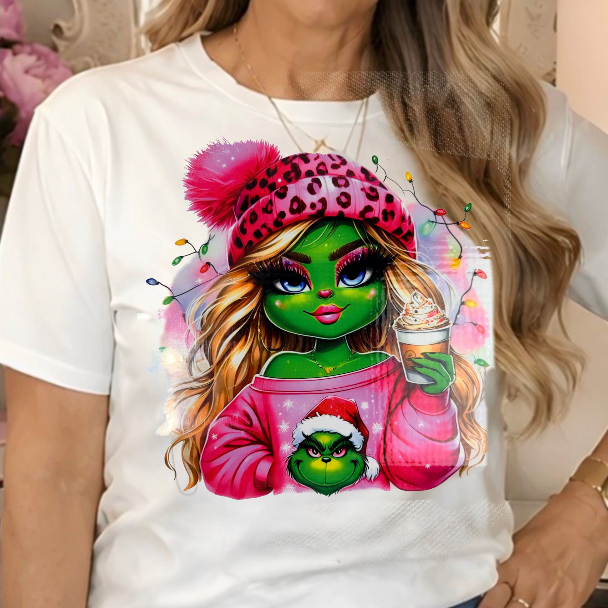 a white graphic t-shirt featuring a colorful illustration of a Sassy Grinchette  in a pink leopard-print beanie and holding a Grinch-themed cup.