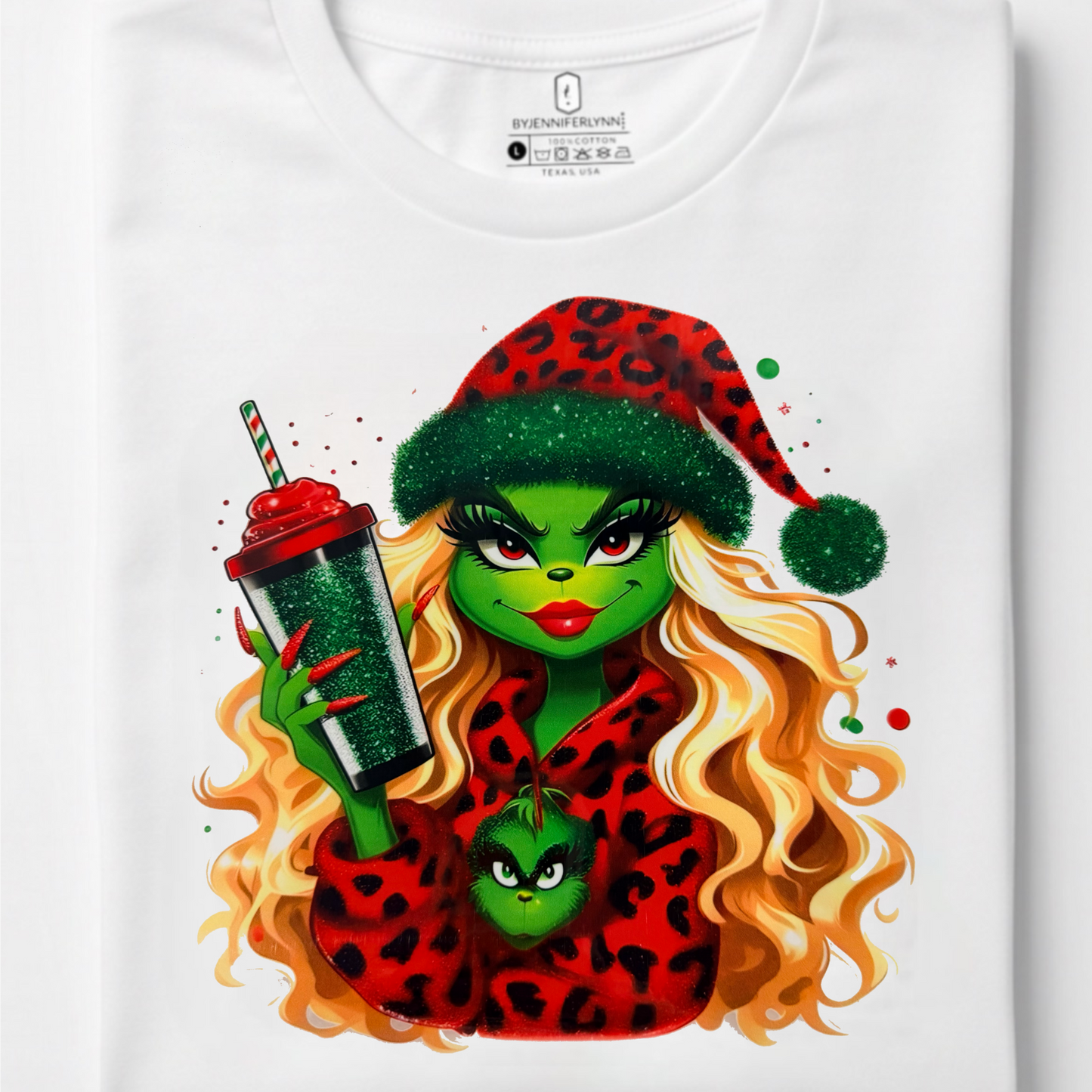 A folded white t-shirt on a light gray background, featuring a colorful graphic print of a green-skinned woman with long blonde hair, wearing a red leopard-print Santa hat and matching robe, holding a green drink.