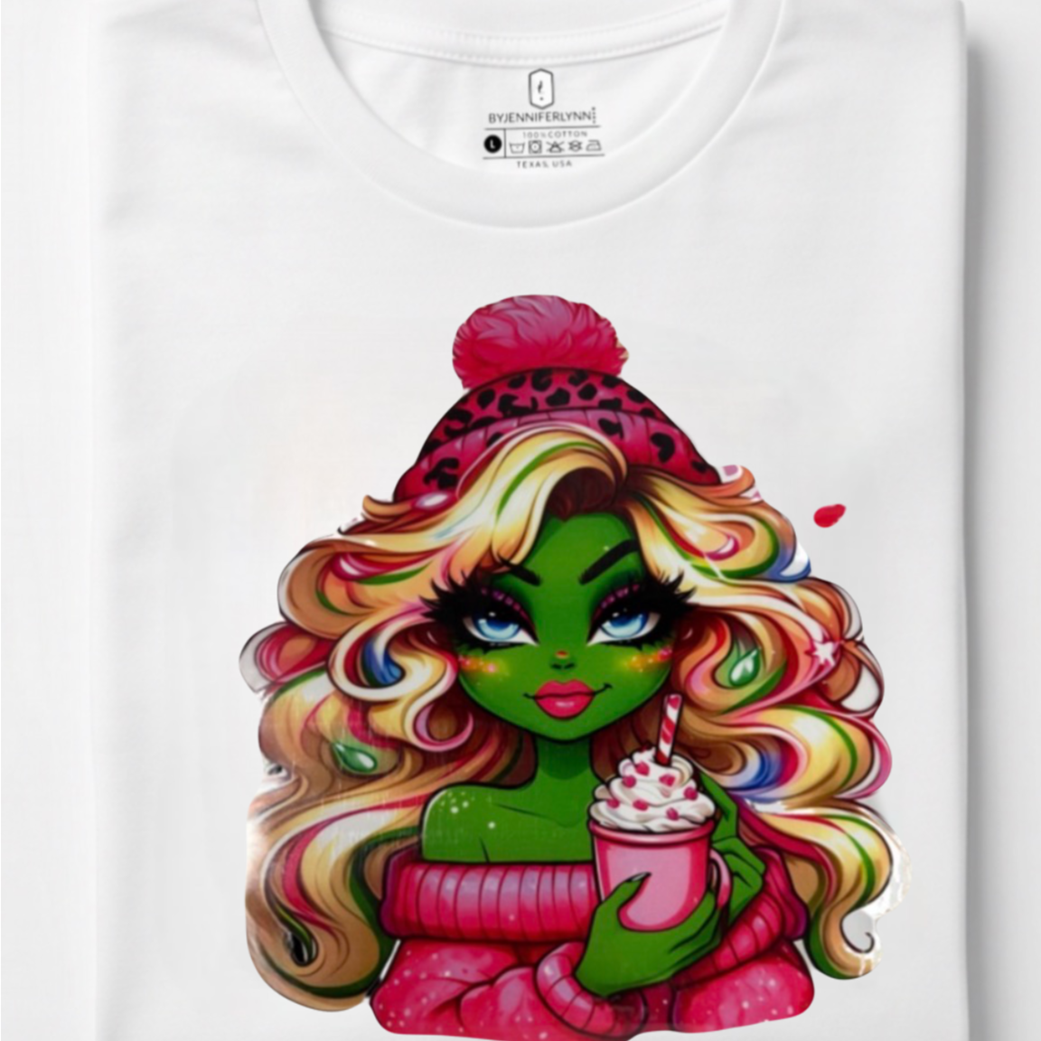 A folded white crewneck t-shirt with a colorful graphic print of a green-skinned woman with long, wavy rainbow hair, wearing a pink beanie and holding a pink drink, displayed on a clean white background.