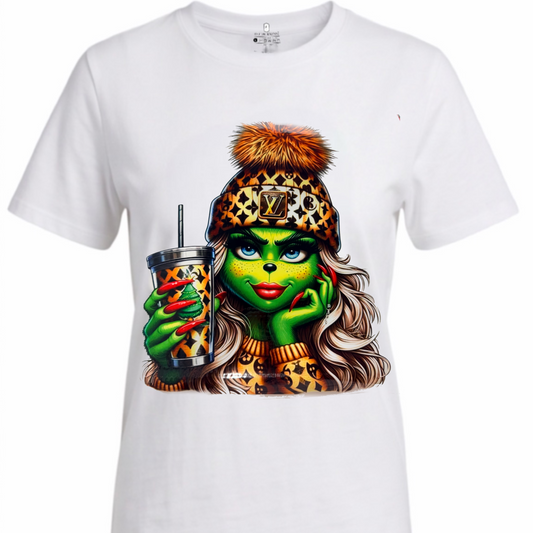 A white short-sleeve crewneck t-shirt featuring a colorful graphic print of a female Grinch woman with leopard-print clothing and a fur-trimmed beanie, holding a drink.