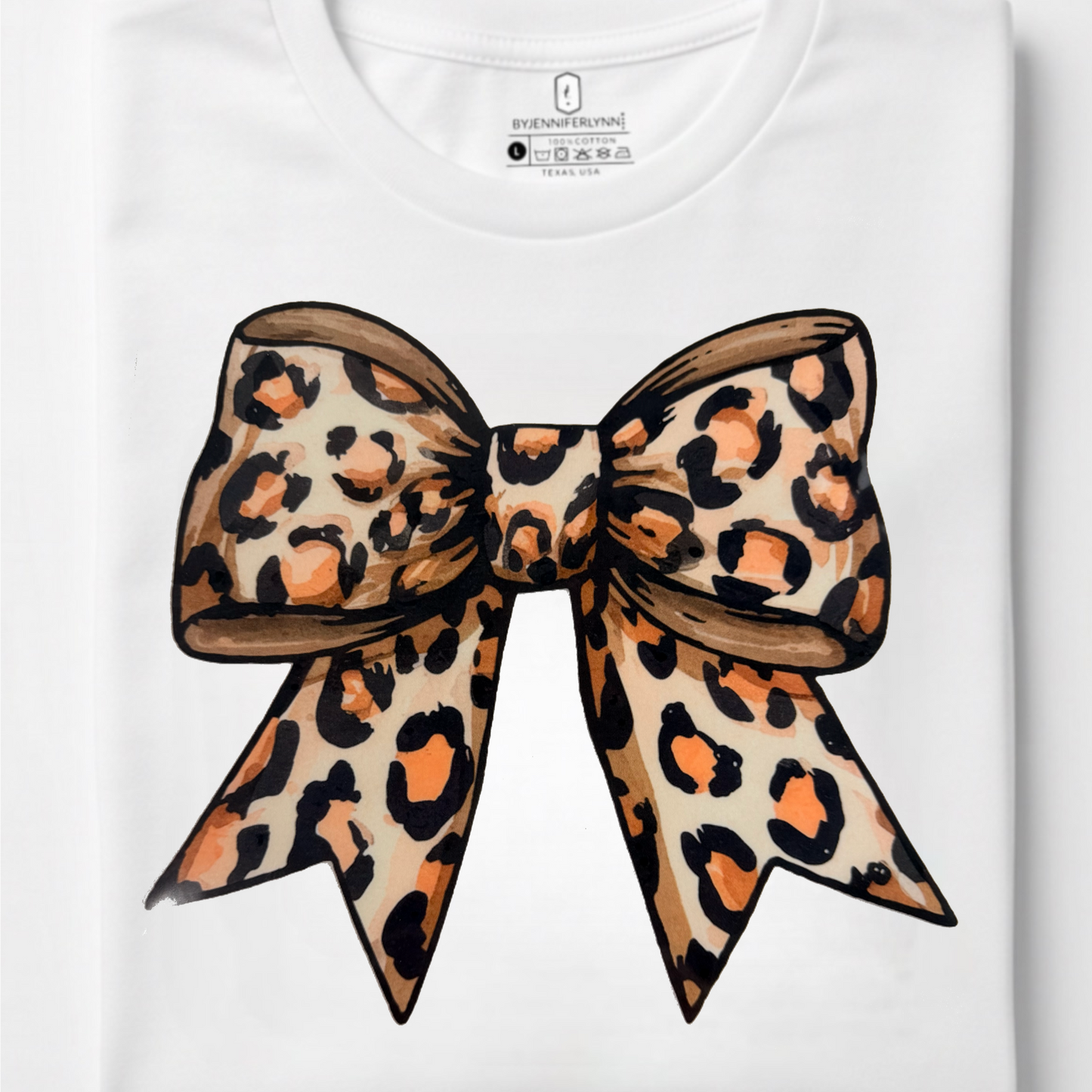 A folded white crewneck t-shirt featuring a large graphic print of a leopard-print bow in beige, brown, and black on the front, set against a clean white background.