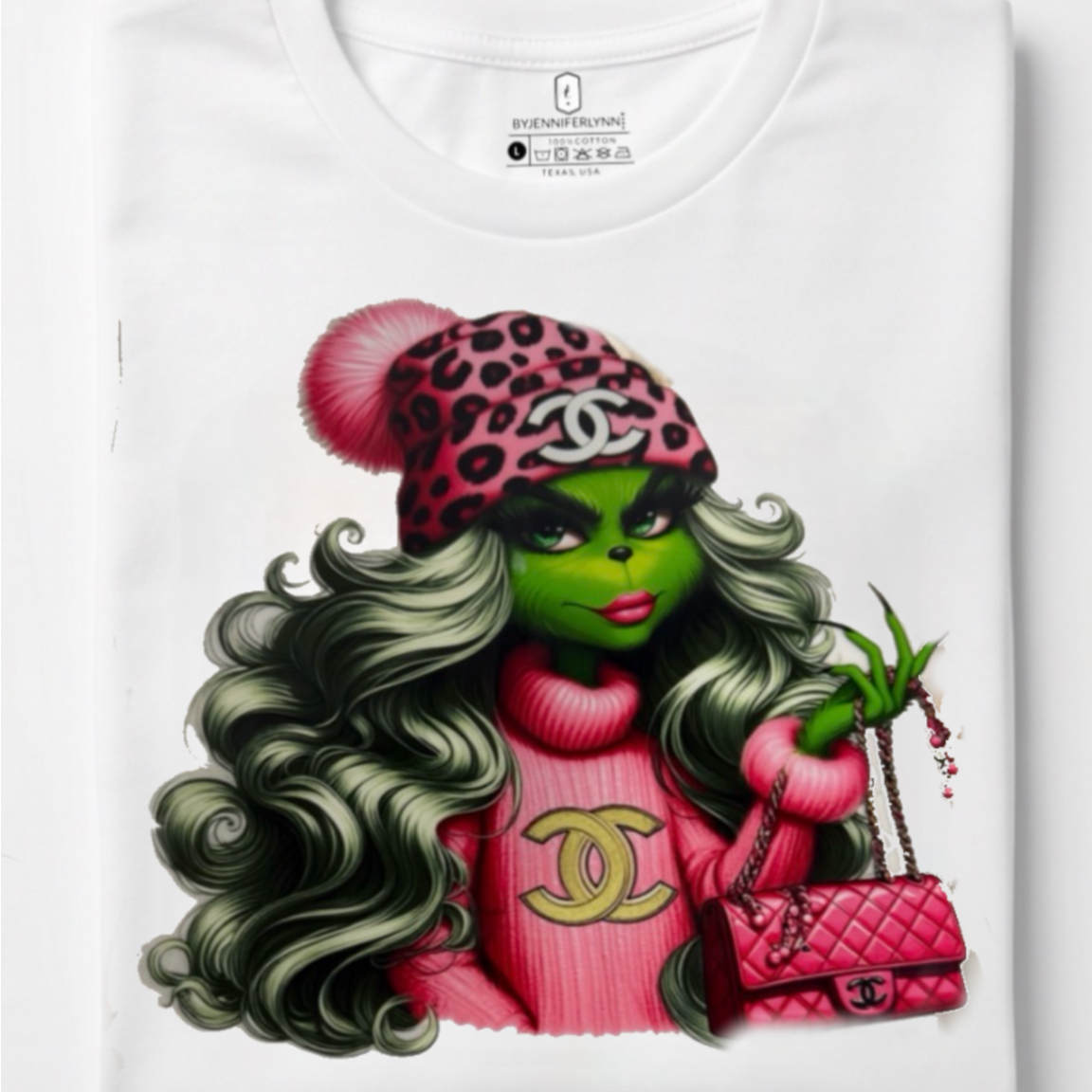 A folded white t-shirt featuring a graphic print of a female Grinch character styled in a pink leopard-print beanie, pink sweater with a gold Chanel logo, holding a pink quilted handbag.