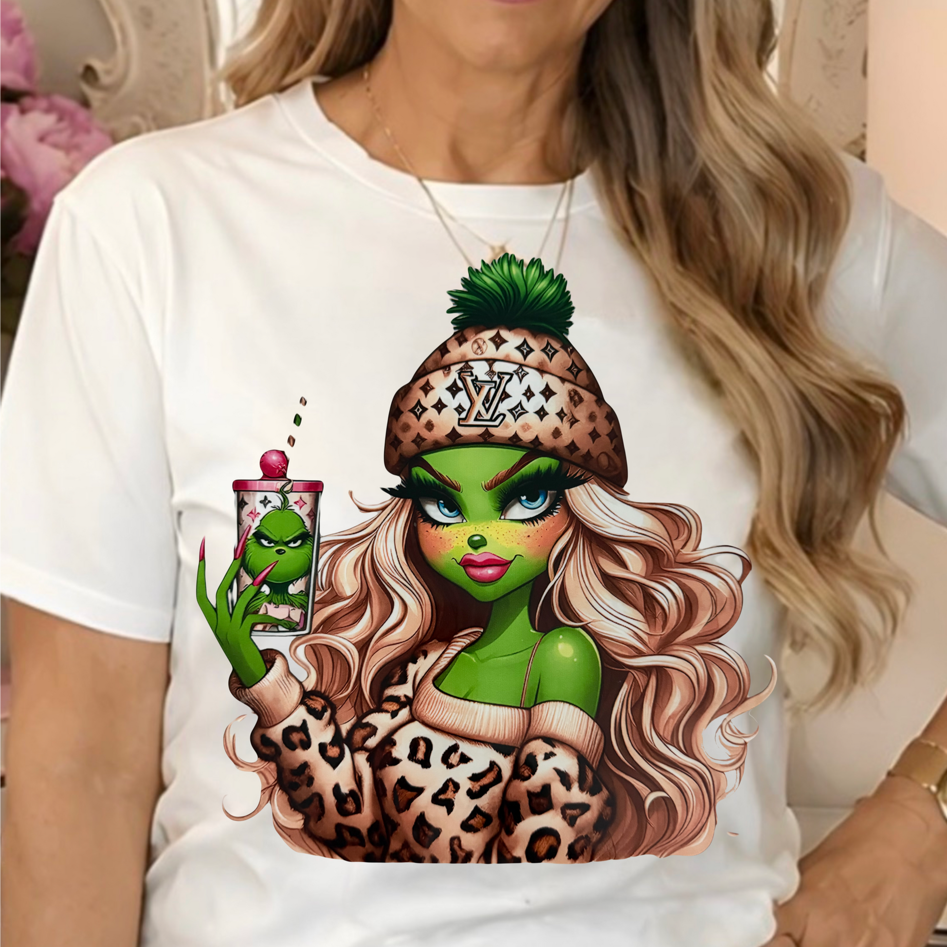 a white graphic t-shirt with a cartoon illustration of a female Grinch in a leopard-print beanie holding a drink.