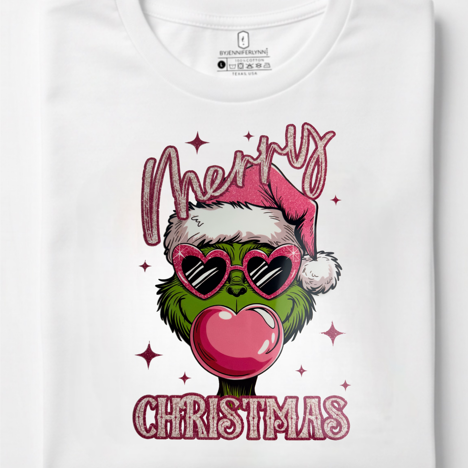 A folded white t-shirt with a colorful graphic of a Grinch character wearing pink heart-shaped sunglasses, a Santa hat, and blowing a large pink bubblegum bubble. The text 'Merry' is above and 'CHRISTMAS' is below in a pink glitter-style font, with small pink stars scattered around.