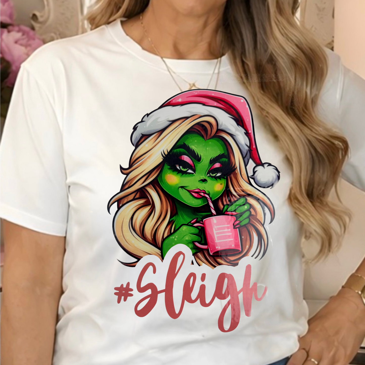a white graphic t-shirt with a cartoon Grinchette wearing a Santa hat and holding a pink drink, with the text '#Sleigh' printed below.