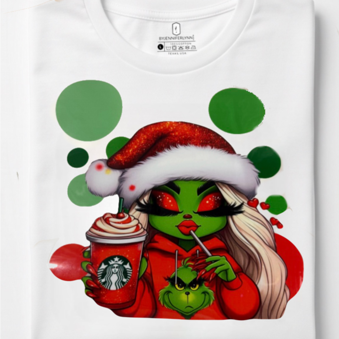 A folded white t-shirt featuring a colorful graphic print of the Grinchette dressed in a Santa hat, holding a Starbucks coffee cup and a phone, with green polka dots in the background.