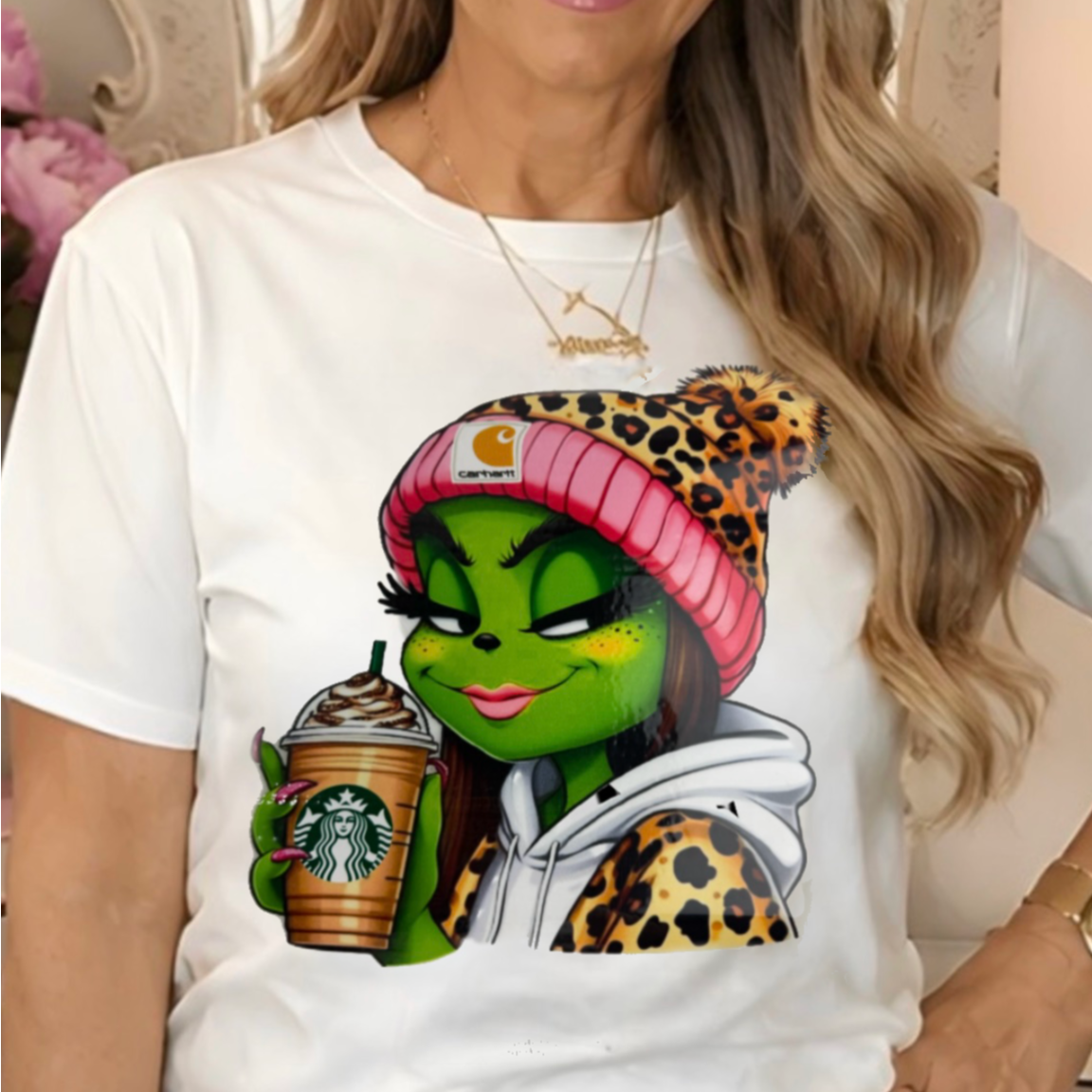 a white graphic t-shirt with a cartoon Grinch character dressed in a pink beanie and leopard print hoodie, holding a Starbucks coffee cup.