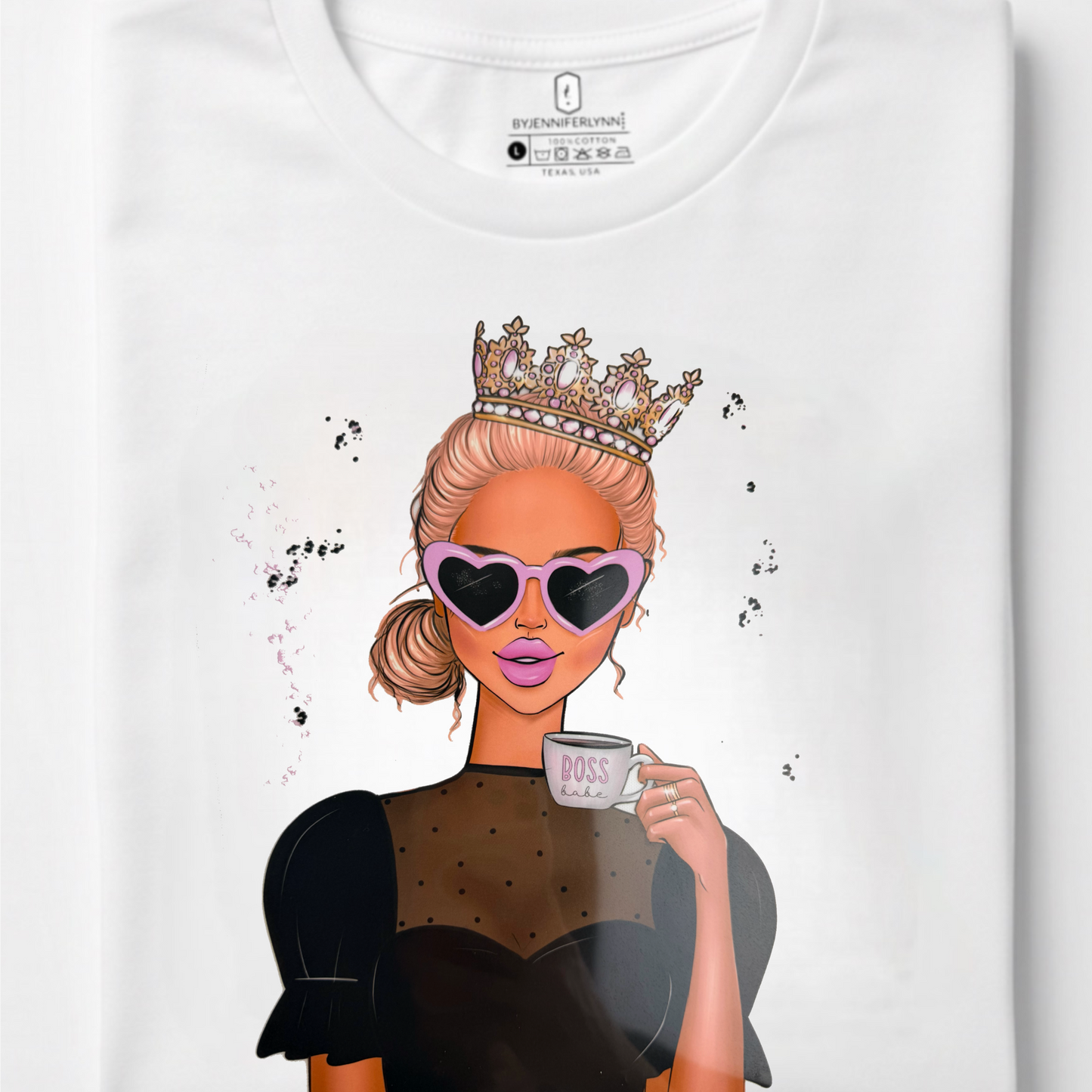 A folded white crewneck t-shirt with a graphic print of a blonde woman wearing a crown, heart-shaped sunglasses, and a polka-dot top, holding a mug.