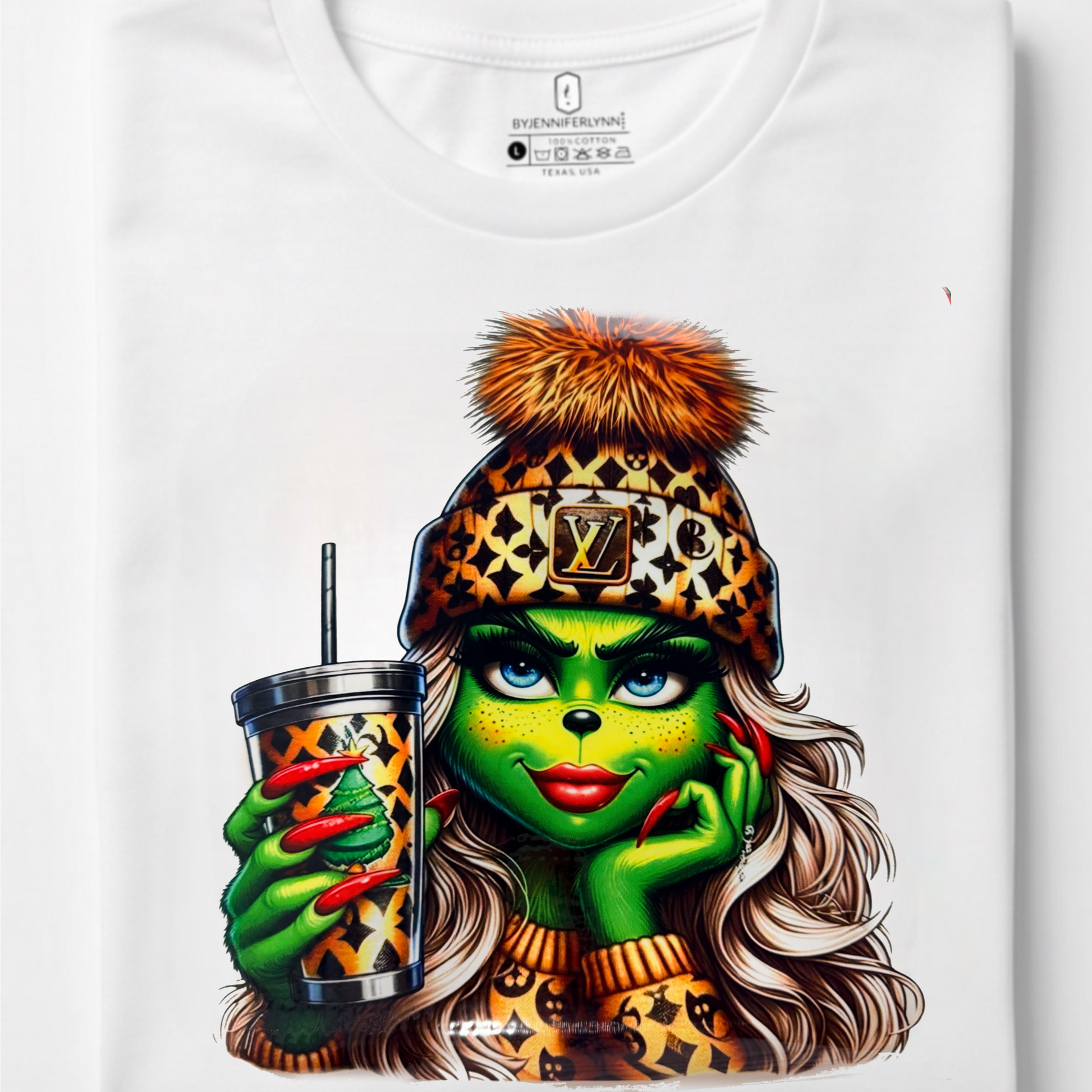 A folded white t-shirt with a graphic print of a female Grinch character wearing a leopard-print beanie with a 'LV' monogram and holding a tumbler.