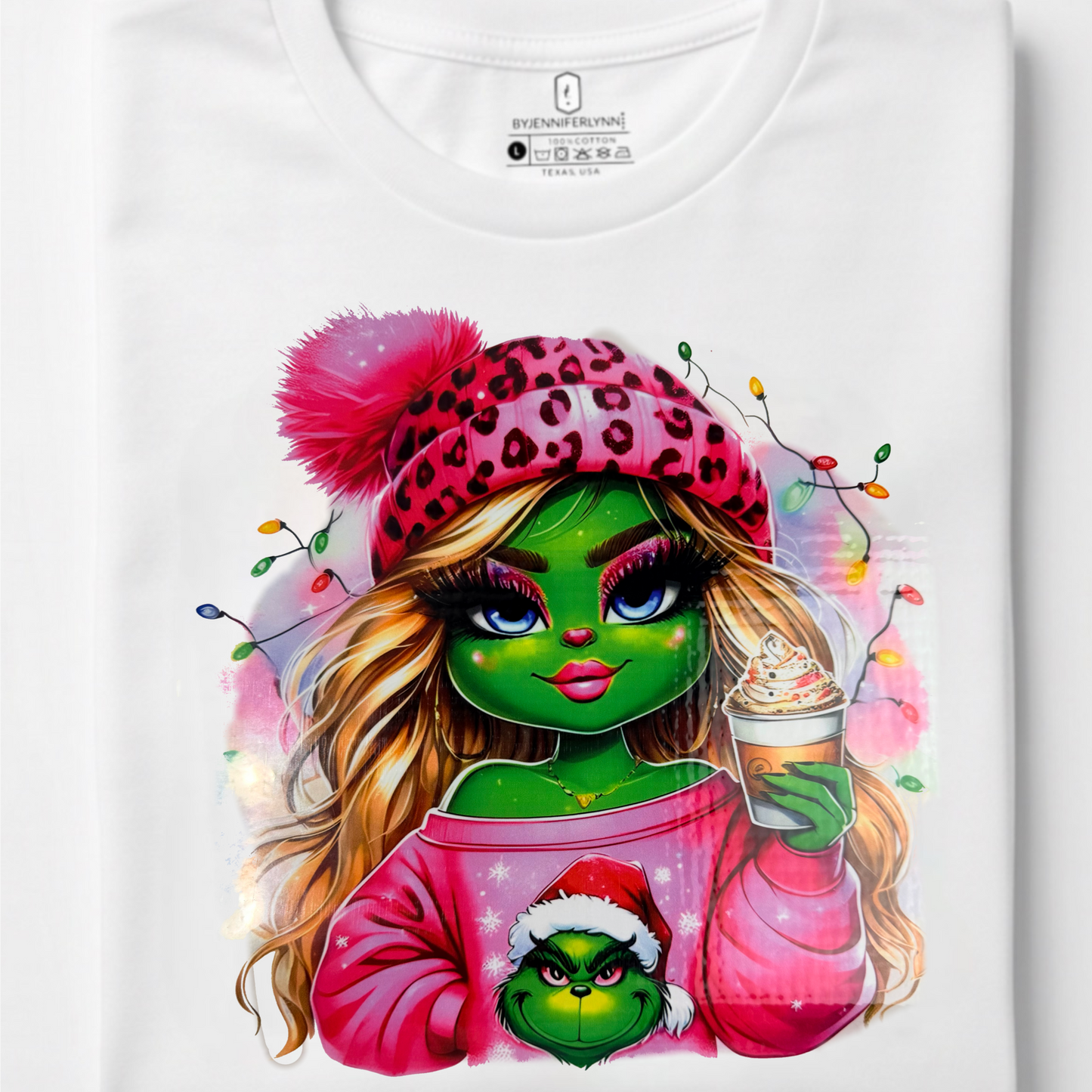 A folded white t-shirt on a light gray background, featuring a colorful graphic print of a green-skinned woman in a pink leopard-print hat and sweater holding a Grinch-themed plush, with Christmas lights and a hot chocolate cup.
