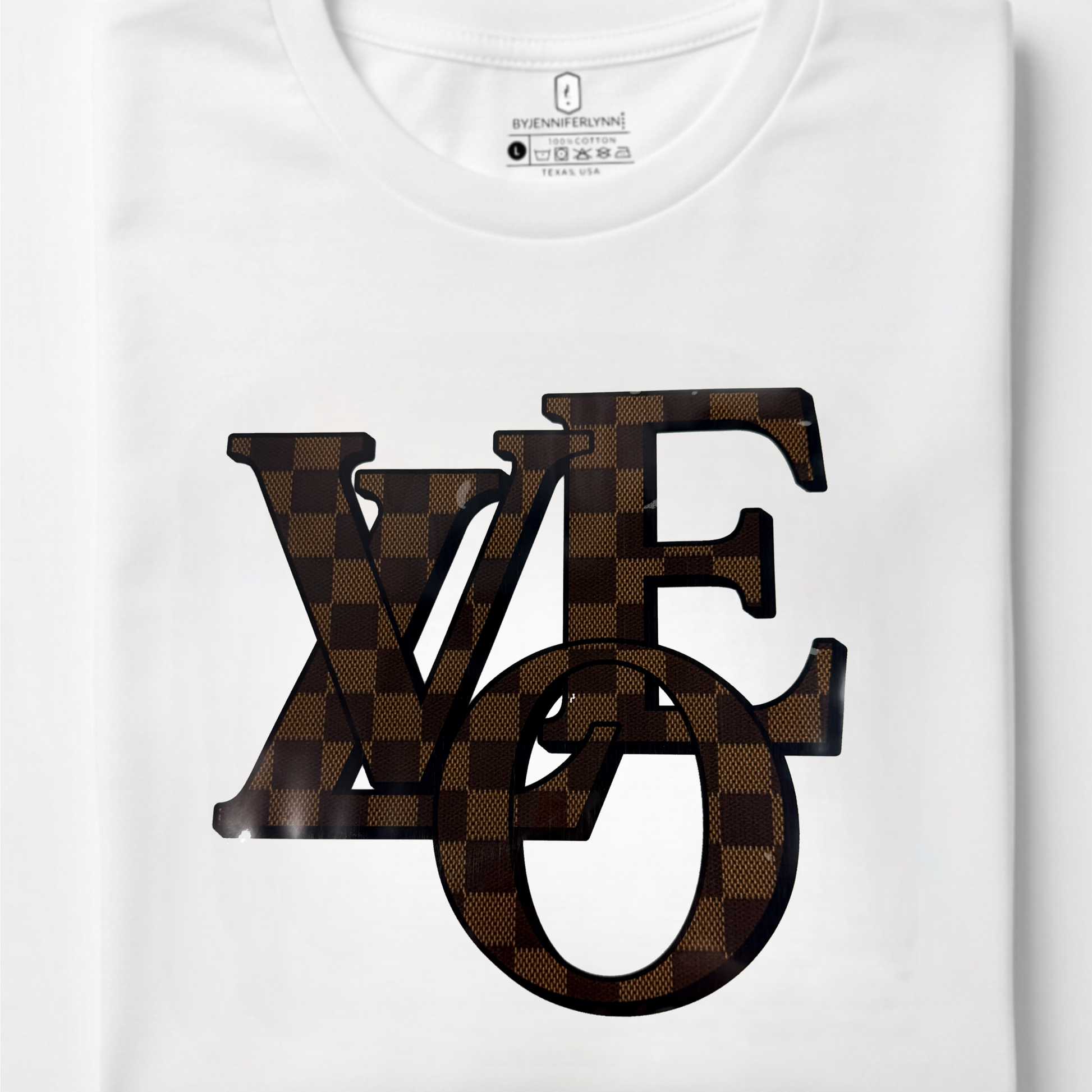 A folded white crewneck t-shirt featuring a large graphic print of the letters 'LOVE' in a brown checkerboard pattern, with 'LOVE' stylized as the classic Louis Vuitton monogram. The neck label clearly shows the Louis Vuitton brand logo.