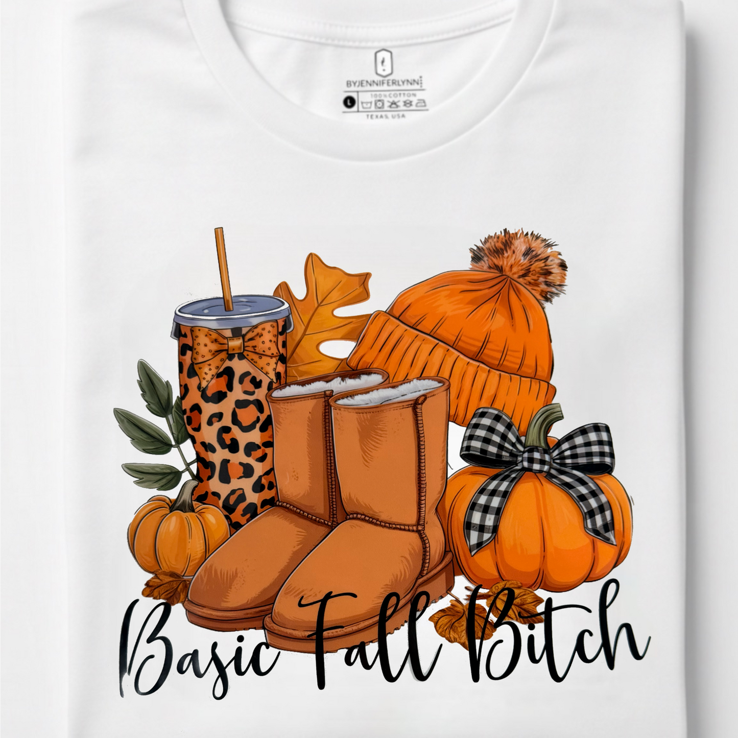 A folded white t-shirt with a graphic print of autumn elements including pumpkins, a beanie, boots, and a leopard-print cup, with the text 'Basic Fall Bitch' printed below.