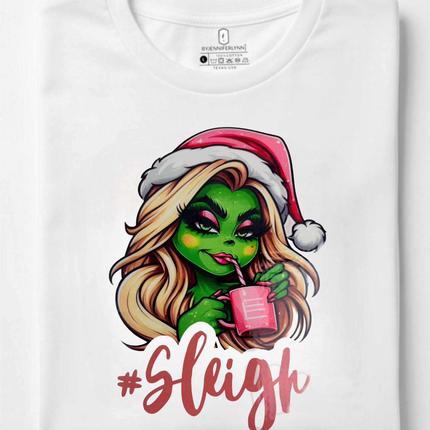A folded white crewneck t-shirt featuring a colorful graphic of a green-skinned woman with blonde hair wearing a red Santa hat and holding a pink drink, with the text '#Sleigh' printed below in red.